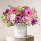 A vibrant flower arrangement featuring pink and purple gerbera daisies, pink and light purple roses, green hydrangeas, and eucalyptus leaves, presented in a white cylindrical box labeled 'BLOOM MAGIC' and displayed on a stone pedestal against a textured beige wall.