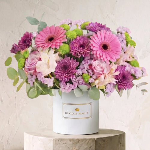 A vibrant flower arrangement featuring pink and purple gerbera daisies, pink and light purple roses, green hydrangeas, and eucalyptus leaves, presented in a white cylindrical box labeled 'BLOOM MAGIC' and displayed on a stone pedestal against a textured beige wall.