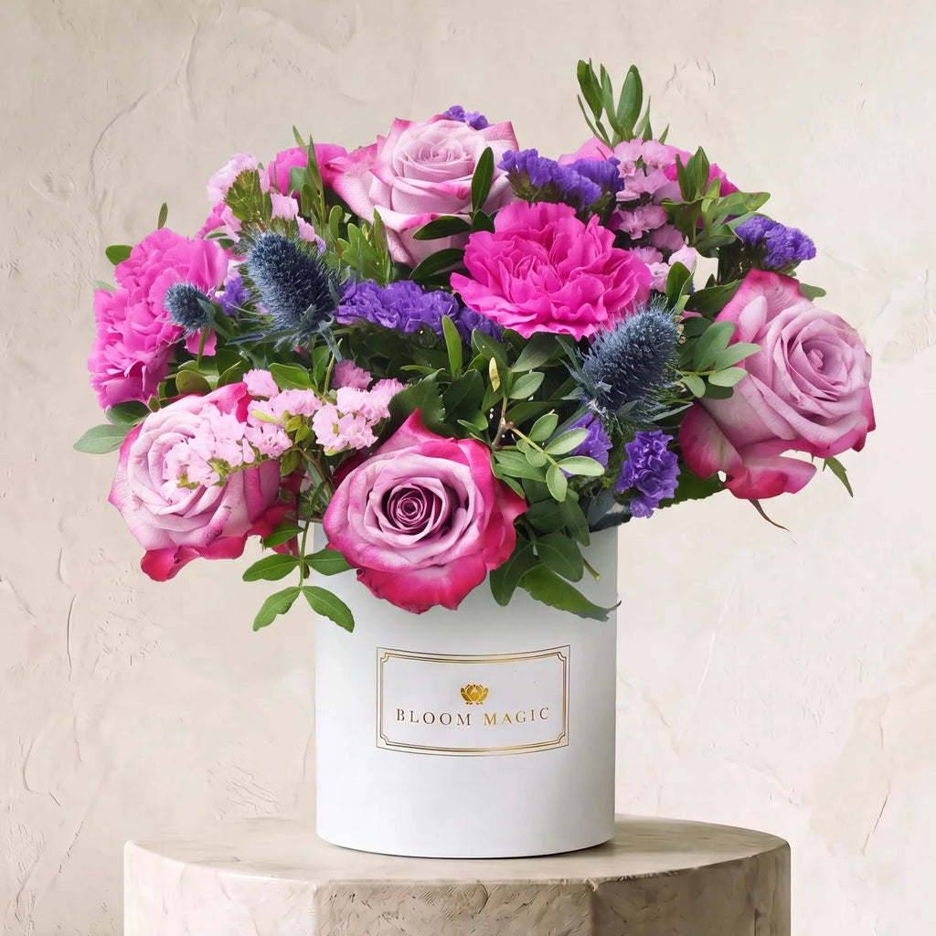 A vibrant floral arrangement featuring pink and purple roses, blue thistles, and green foliage, presented in a white cylindrical box labeled 'BLOOM MAGIC' and displayed on a stone pedestal against a textured beige wall.