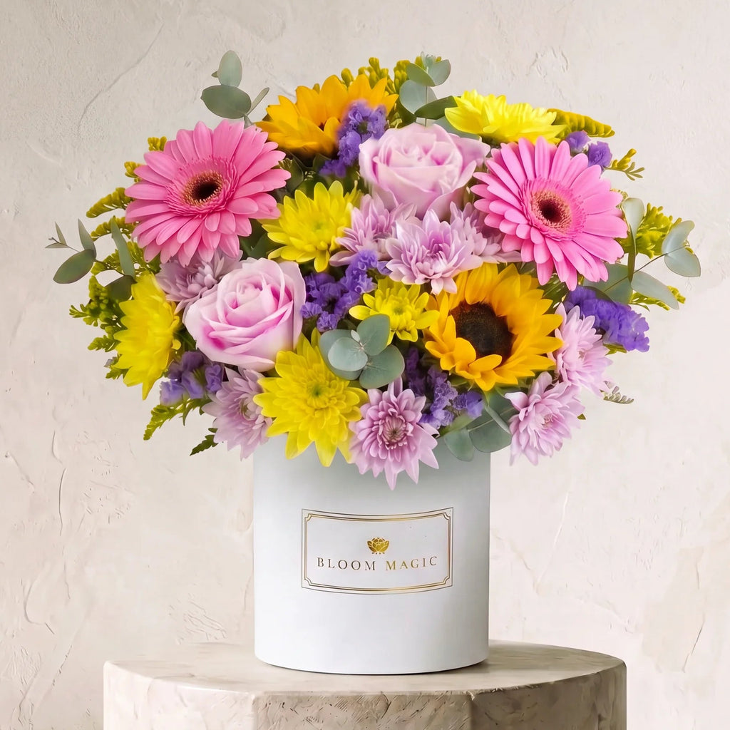 A vibrant flower bouquet featuring pink gerbera daisies, pink roses, yellow sunflowers, purple asters, and green eucalyptus stems, presented in a white cylindrical pot labeled 'BLOOM MAGIC' and displayed on a stone pedestal against a textured beige wall