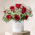 A floral arrangement of red roses and white baby's breath in a white cylindrical box labeled BLOOM MAGIC, displayed on a stone pedestal against a textured beige wall.