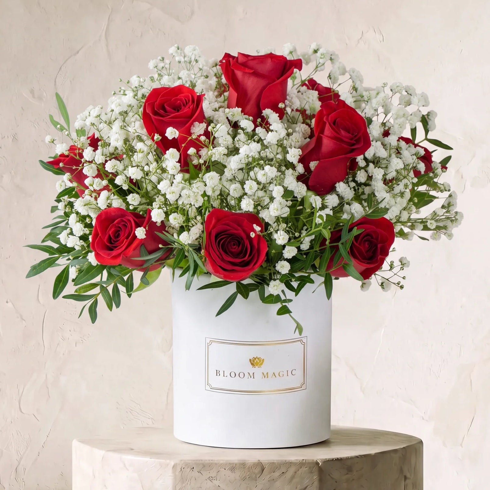 A floral arrangement of red roses and white baby's breath in a white cylindrical box labeled BLOOM MAGIC, displayed on a stone pedestal against a textured beige wall.